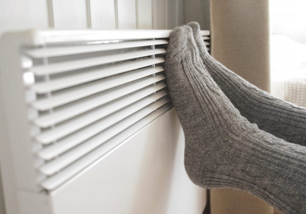 Legs in knitted socks warming over electric heater. Frozen woman wearing a warm woolen socks freezing for winter cold. Discomfort spending time at home. Girl warming her feet on modern radiator.